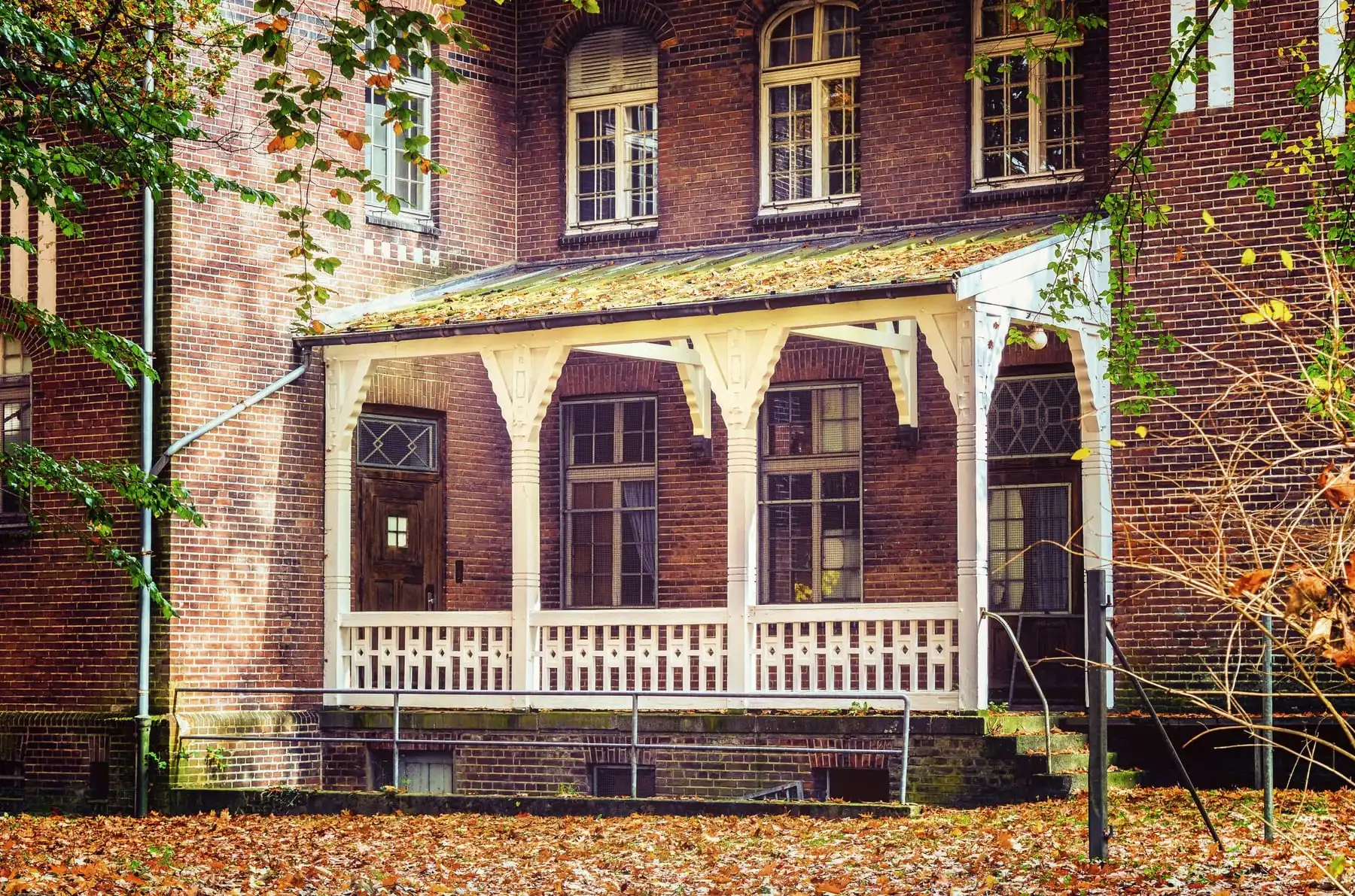 A red-brick family home with a white wooden porch, set among autumn trees