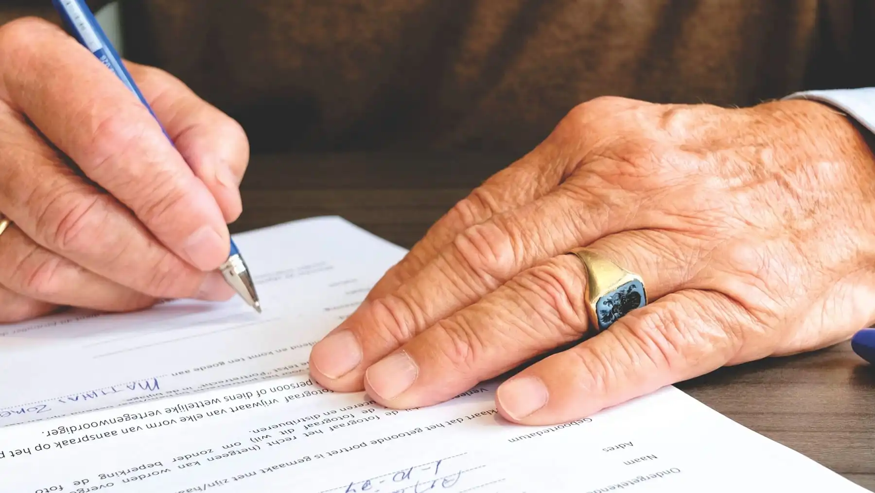 An older person's hands signing a document with a pen, a gold ring on one finger